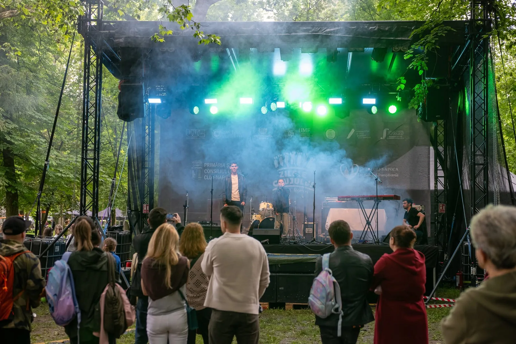 Public în fața unei scene între arbori, cu lumină verde și albă